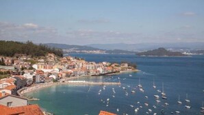 Un poble costaner amb edificis al passeig marítim, una marina plena de vaixells i una vista clara del mar i les muntanyes llunyanes.