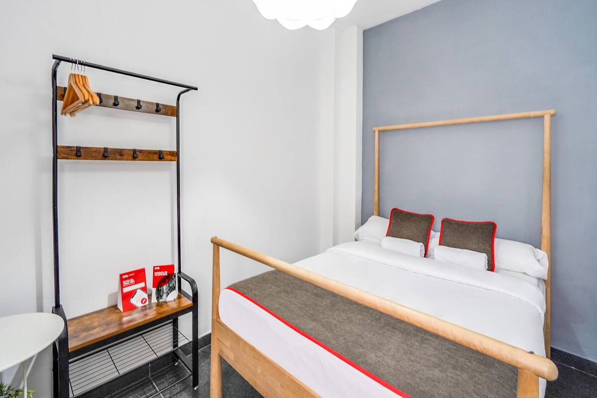 A single bed with a wooden frame and red accents, a metal rack with clothes, and a small table with a red sign.
