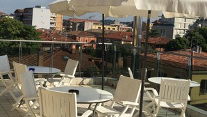A rooftop terrace with white chairs and tables, a large white umbrella, and a view of a cityscape with red-tiled roofs. - Hotel Athena (Pesaro)