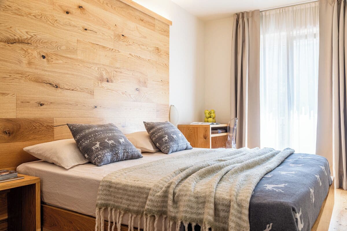 A bedroom with a wooden headboard, a bed with patterned pillows, a bedside table with a lamp, and a window with curtains.