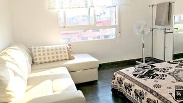 Una habitación con un sofá blanco, una colcha estampada, un ventilador de pie y una ventana con vistas a edificios.