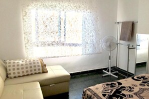Une pièce comprenant un canapé, une couvre-lit à motifs, un ventilateur sur pied et une fenêtre agrémentée de rideaux décoratifs.