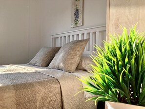 Un lit avec une couette et des oreillers de couleur beige, une tête de lit blanche et une plante en pot placée à droite.