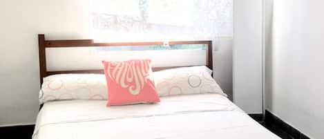A bed with white bedding and a pink pillow, a wooden headboard, and a window with patterned curtains.
