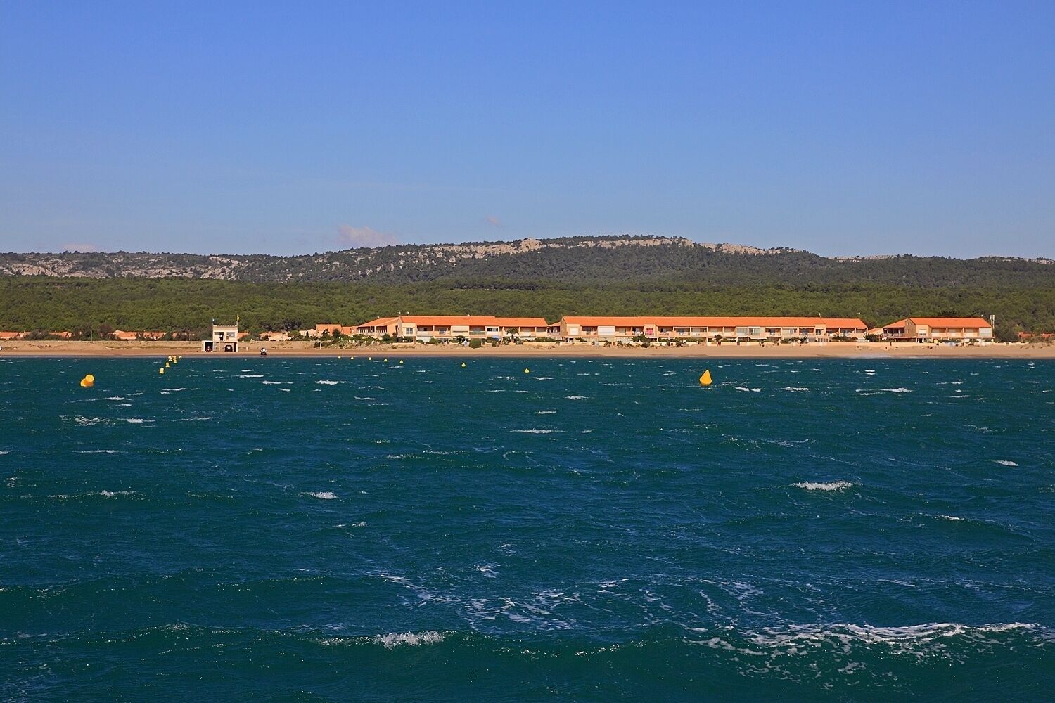 Un hôtel situé sur la côte, composé de plusieurs bâtiments aux toits orange, avec une plage en arrière-plan et des bouées dans l’eau.