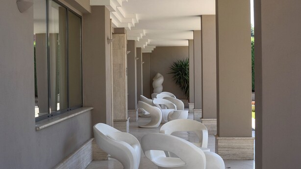 A modern interior with a series of white, sculptural chairs arranged in a row, a large window on the left, and a plant in the background. - Hotel Città dei Papi (Anagni)