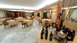 A spacious hotel dining area with multiple round tables, each set with chairs and covered with yellow tablecloths. There are several wine bottles and glasses on a central table, and the floor is made of marble. - Hotel Città dei Papi (Anagni)