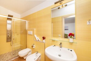 Ein Badezimmer mit gelb gefliesten Wänden, einer Duschkabine aus Glas, einer Toilette, einem Bidet, einem Waschbecken und einem Spiegel.