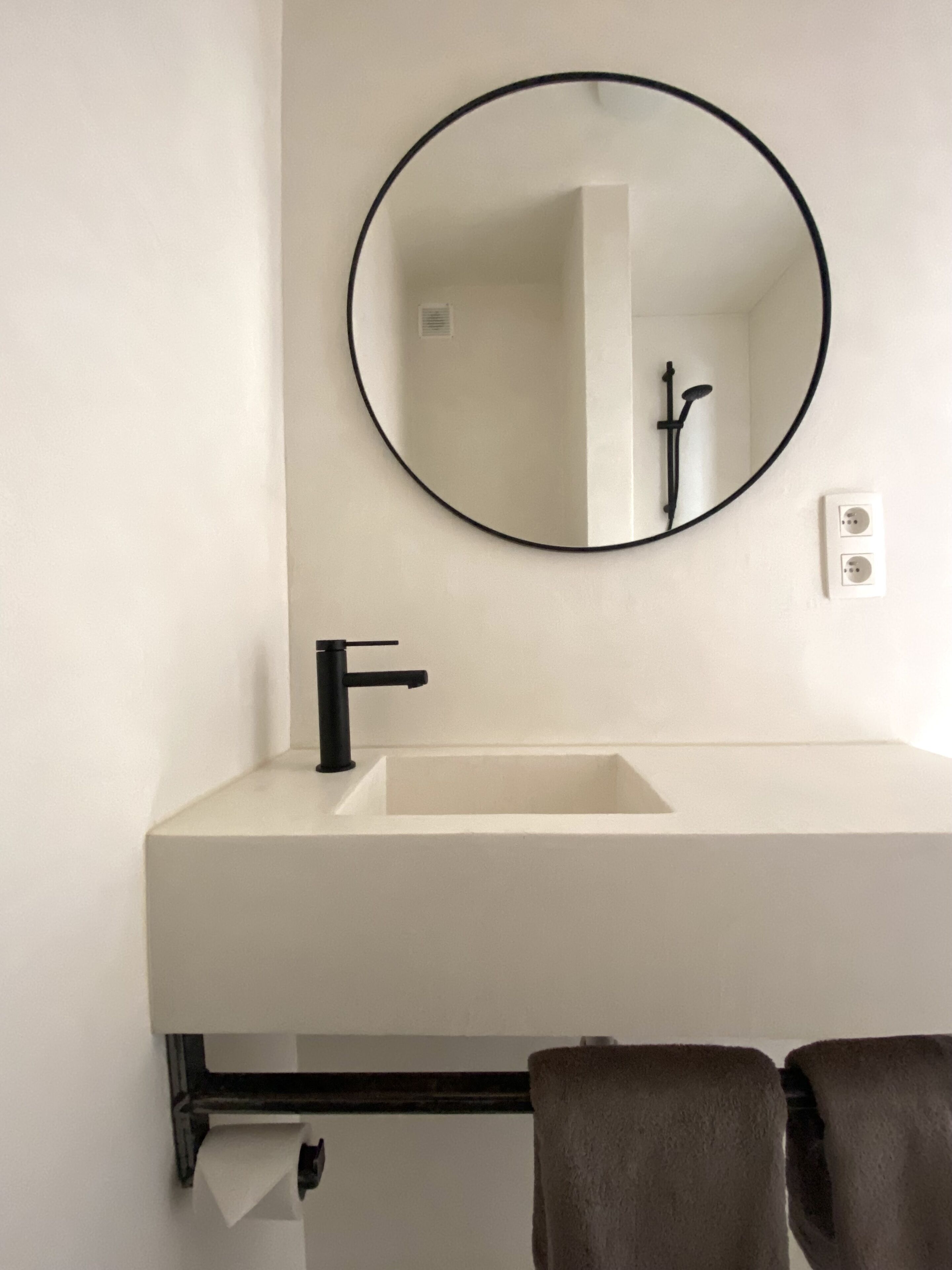 Casa de banho com lavatório branco, torneira preta e espelho redondo. Há um porta-toalhas com duas toalhas e um dispensador de papel.