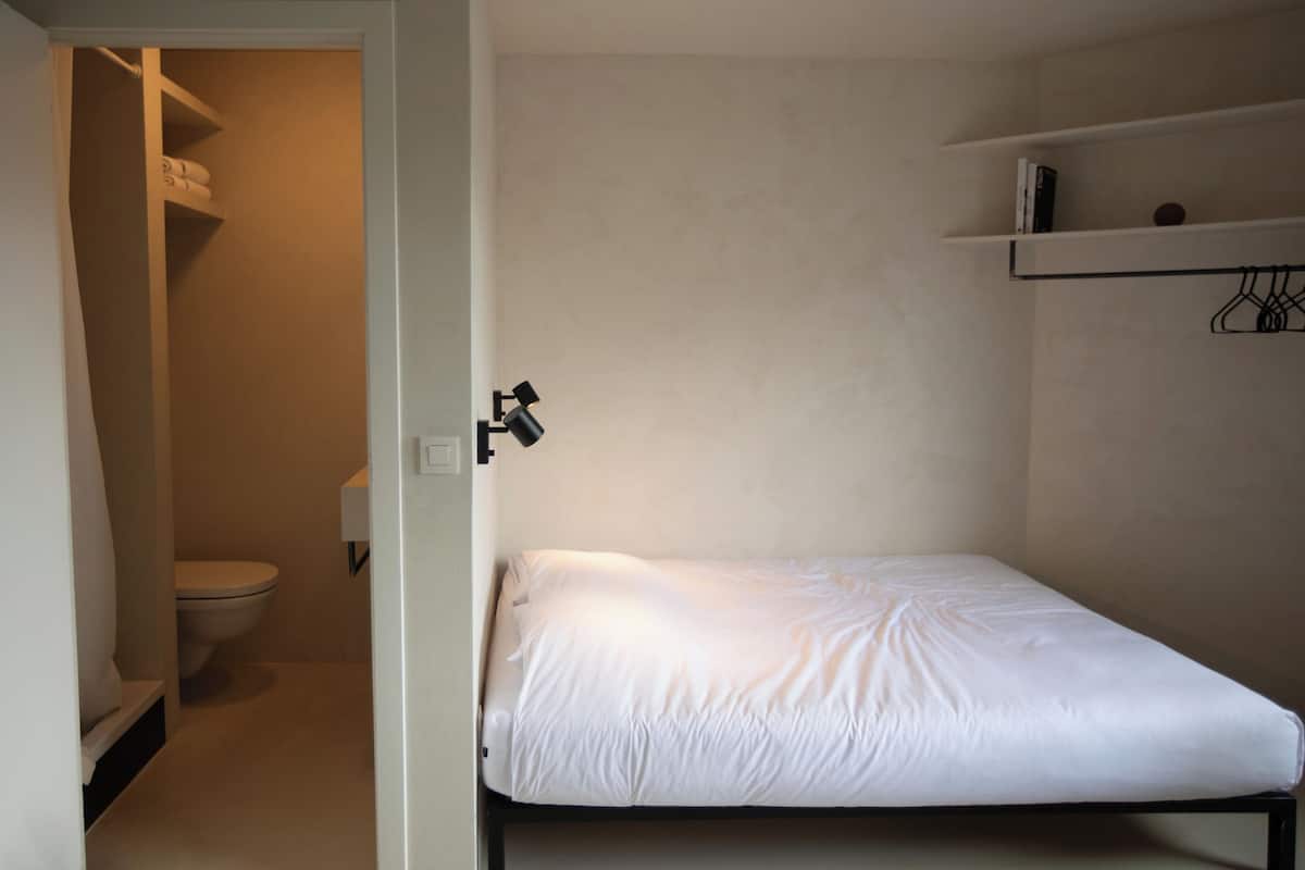A small, single-bed room with a white mattress, a white toilet, a shelf with a book, and a hanging clothes rack.
