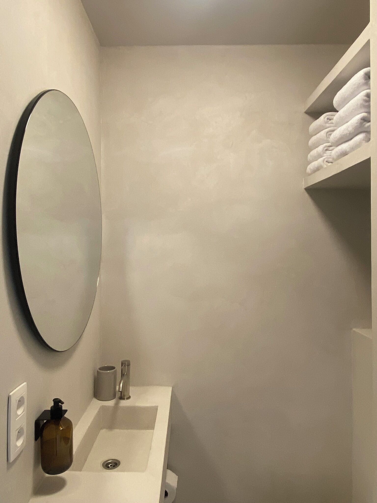 A small bathroom with a white sink, a round mirror, a towel rack, and a soap dispenser.