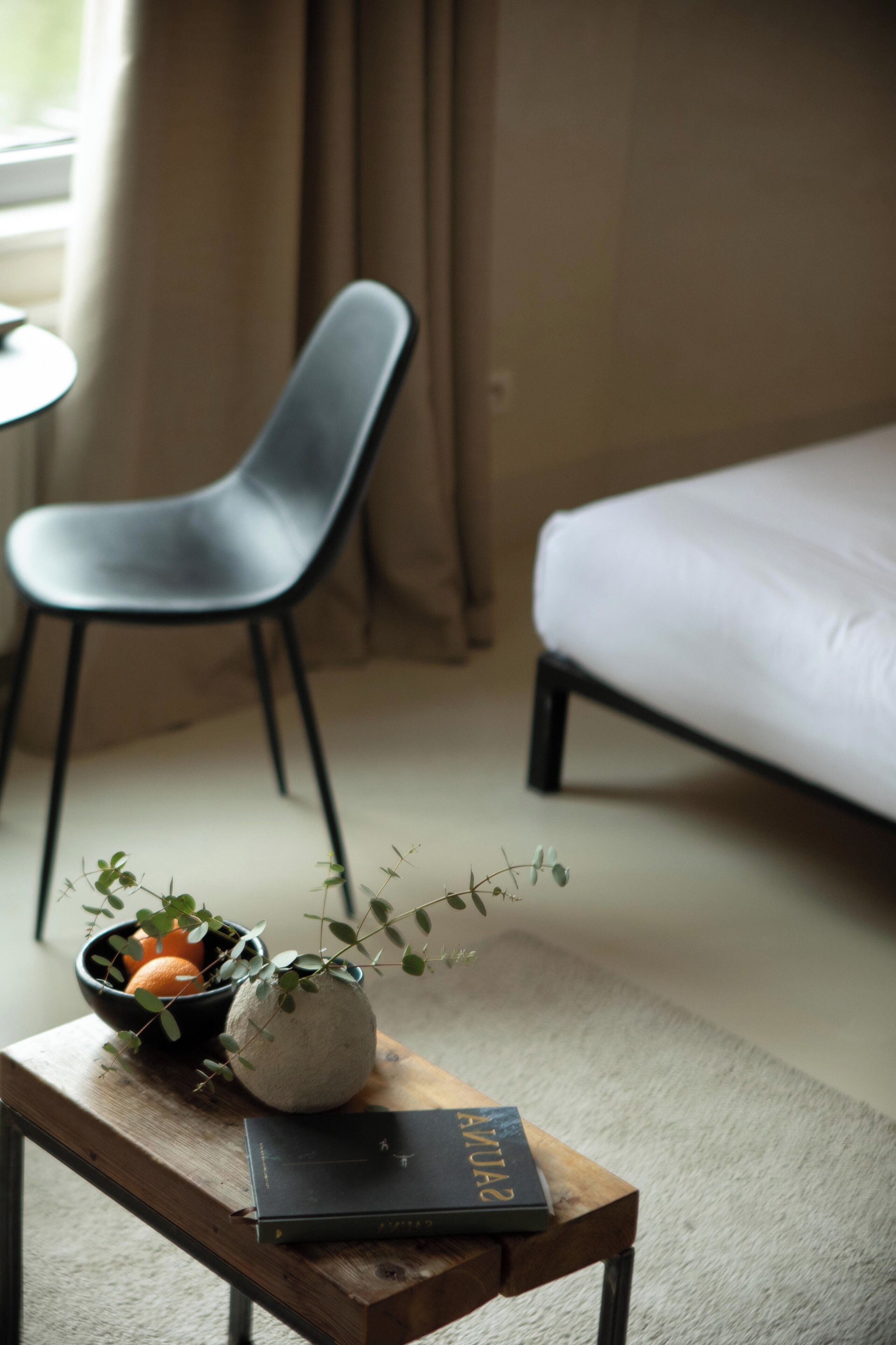 A room with a bed, a chair, a small table with a book and a bowl of fruit, and a window with curtains.