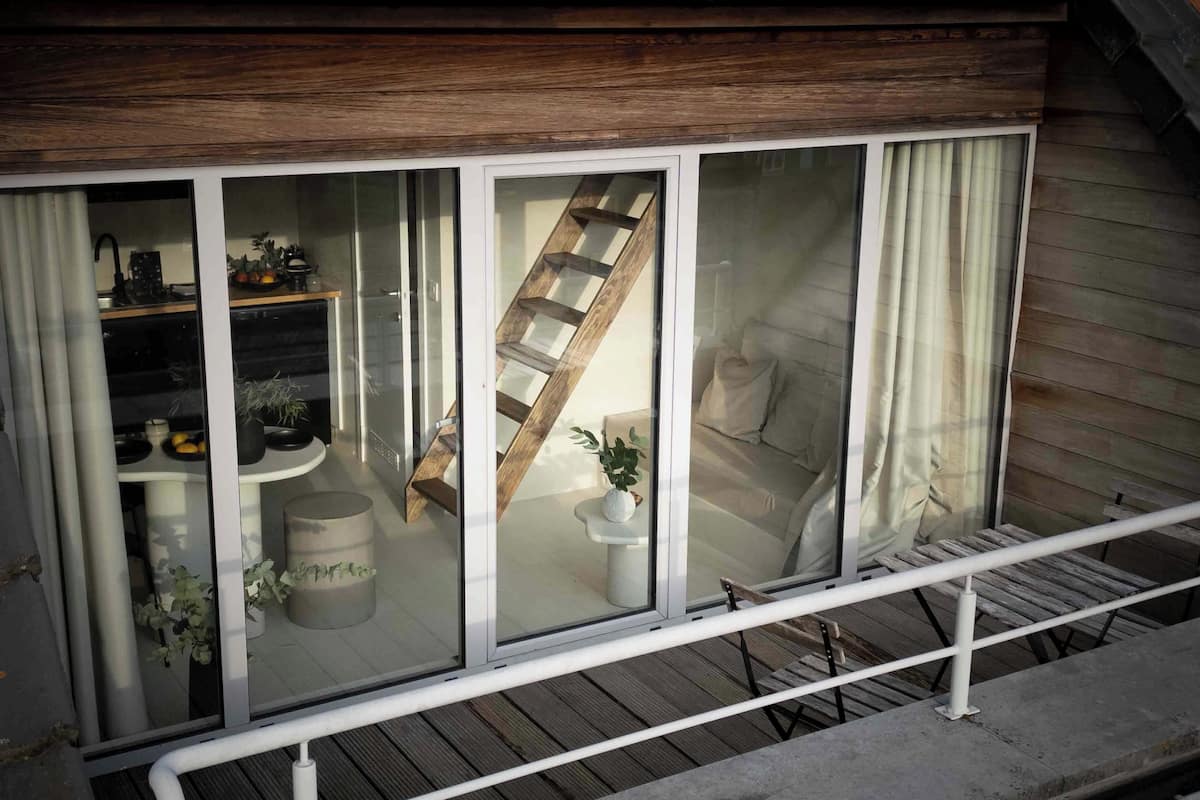 A balcony with a wooden railing, a glass door leading to a room with a bed, a small table with a plant, and a staircase visible through the glass.