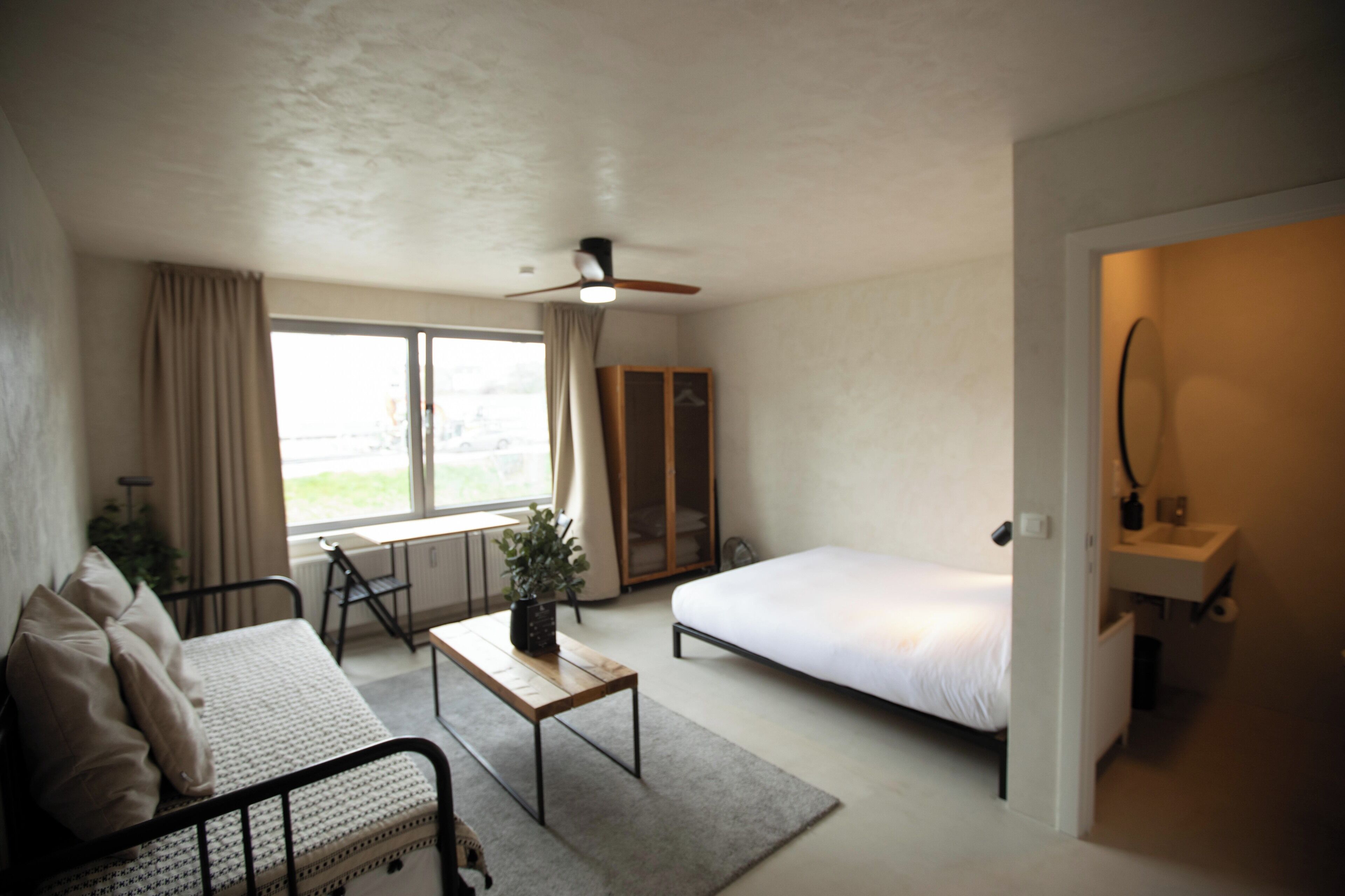 Una habitación de hotel compacta con cama, un escritorio pequeño, una silla, un sofá, un ventilador de techo, una ventana con cortinas y un baño con lavabo y espejo.