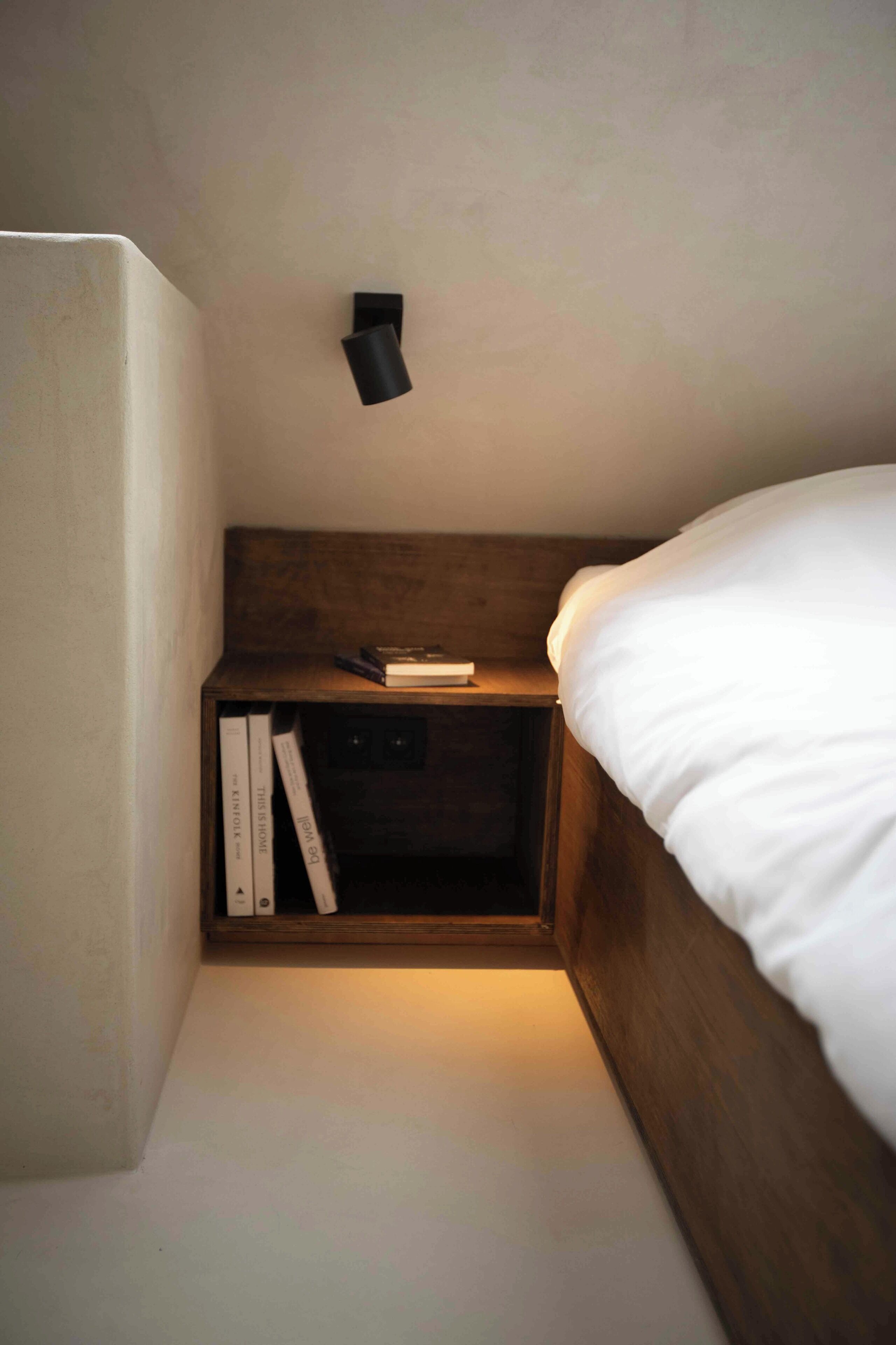 Ein kleines, gemütliches Schlafzimmer mit einem hölzernen Kopfende, einem Nachttisch mit Büchern und einer Wandleuchte.