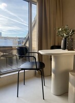 A modern room with a large window, a round table, a black chair, a vase with green leaves, and a sign on the table.