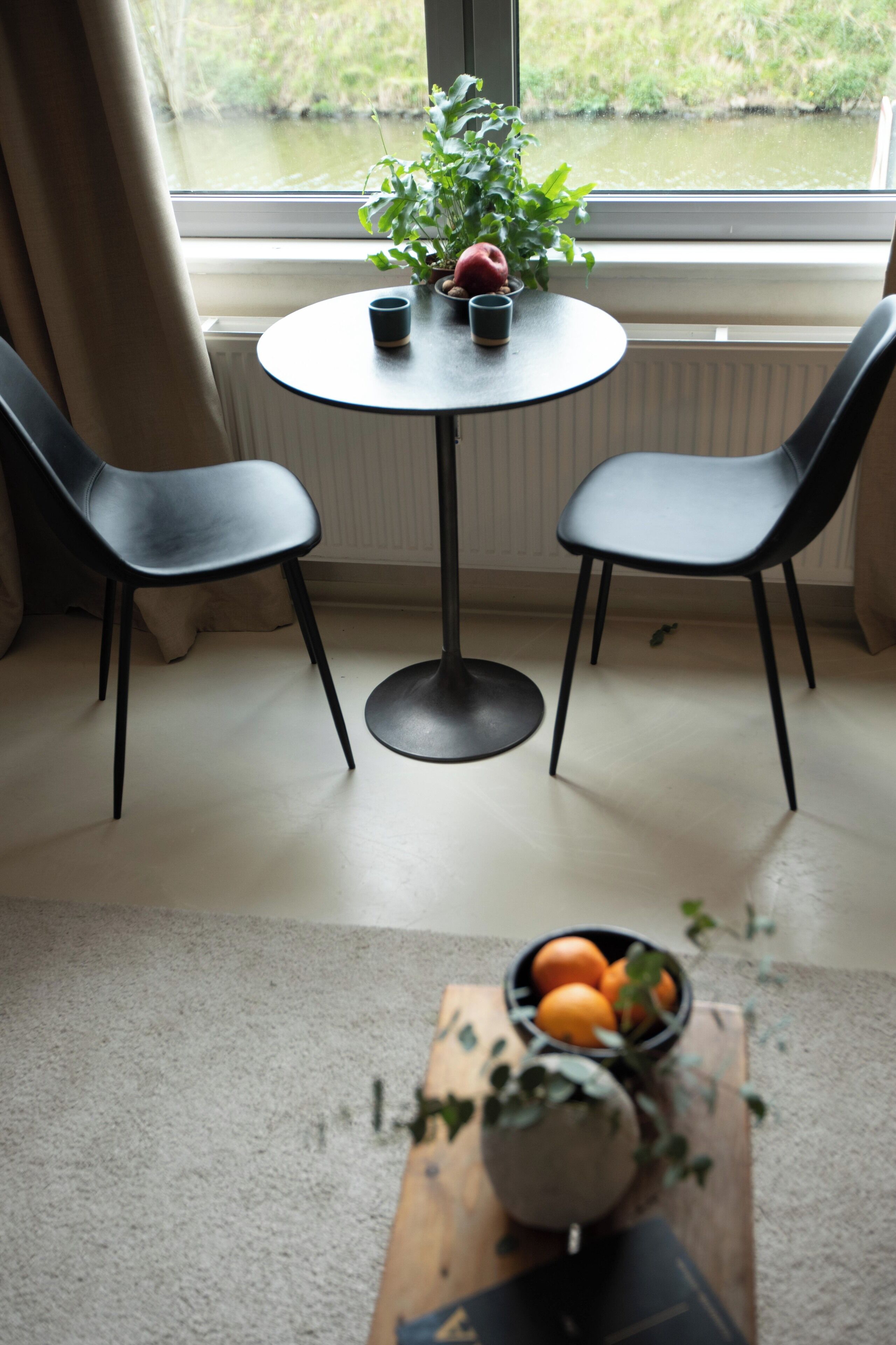 Una habitación con una mesa redonda, dos sillas, una planta en maceta y un cuenco de fruta sobre una mesa de madera.