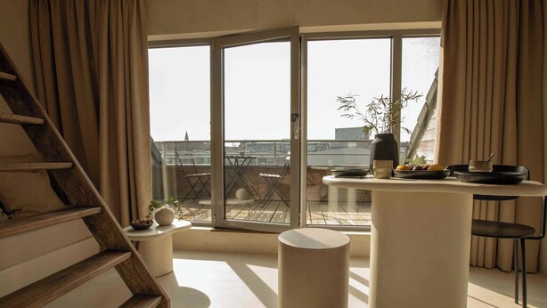 A modern living space with a wooden staircase, a round table, and a view of the city through large windows.