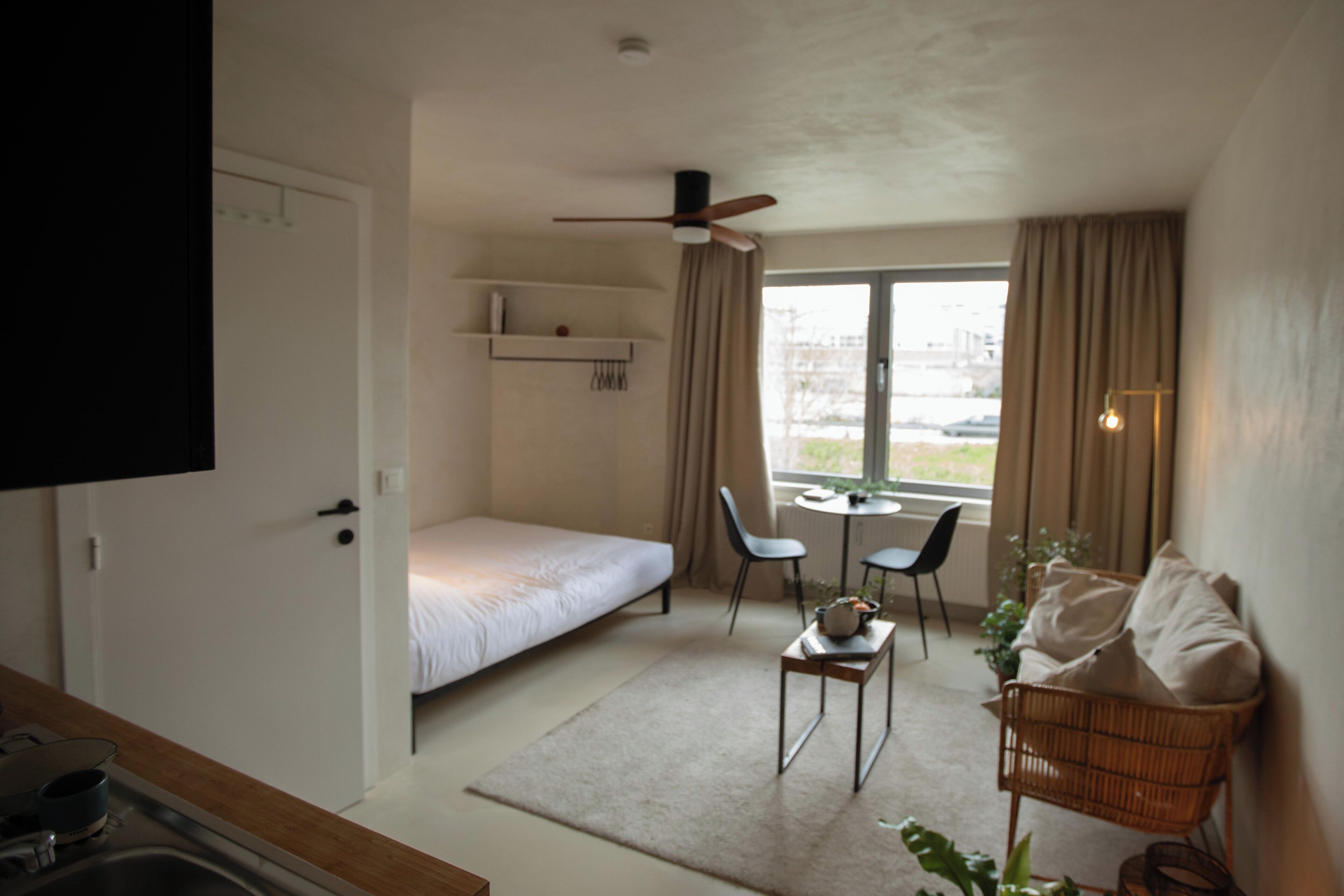 Quarto com cama, escrivaninha, cadeiras, mesa pequena, ventilador de teto e janela com cortinas.