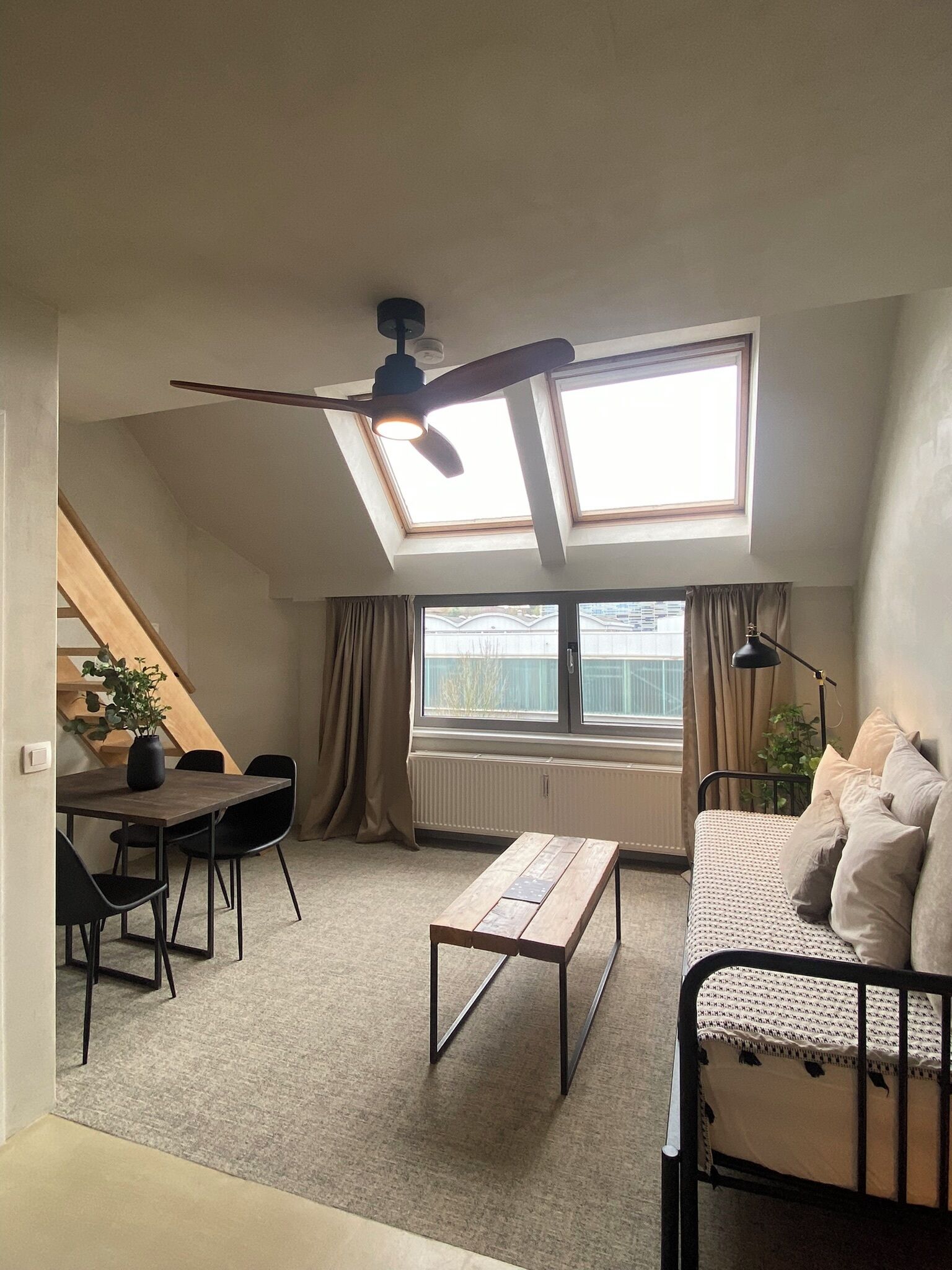 A loft-style room with a sloped ceiling, skylight windows, a dining area with a table and chairs, a sofa, and a small wooden bench.