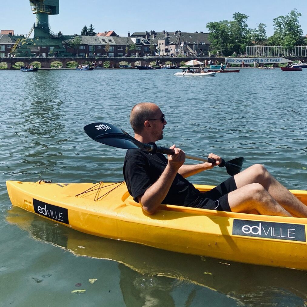 Una persona en un kayak amarillo con la marca 'edville', remando en un río tranquilo con edificios al fondo.