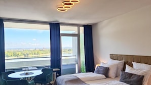 A hotel room with a bed, a table, and chairs, a large window with a view of a lake and greenery, and a modern ceiling light fixture. - Rheinblick Appartments (Cologne)