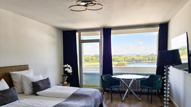 A bedroom with a bed, a desk with chairs, a lamp, and a view of a lake. - Rheinblick Appartments (Cologne)