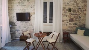 A cozy room with a stone wall, a round wooden table with two chairs, a TV mounted on the wall, and a window with white curtains. - Apartments and Rooms Djanovic (Split)
