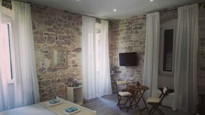 A room with two beds, a small table with chairs, a TV mounted on the wall, and a stone wall. - Apartments and Rooms Djanovic (Split)