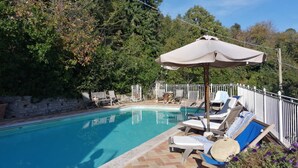 Ein Pool mit Liegestühlen und Sonnenschirm, umgeben von Bäumen und einer Steinmauer.