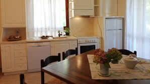 Eine kleine Küche mit weißen Schränken, einem Herd, einem Spülbecken und einem Kühlschrank. Ein Esstisch mit Stühlen und eine Blumenvase stehen ebenfalls bereit.