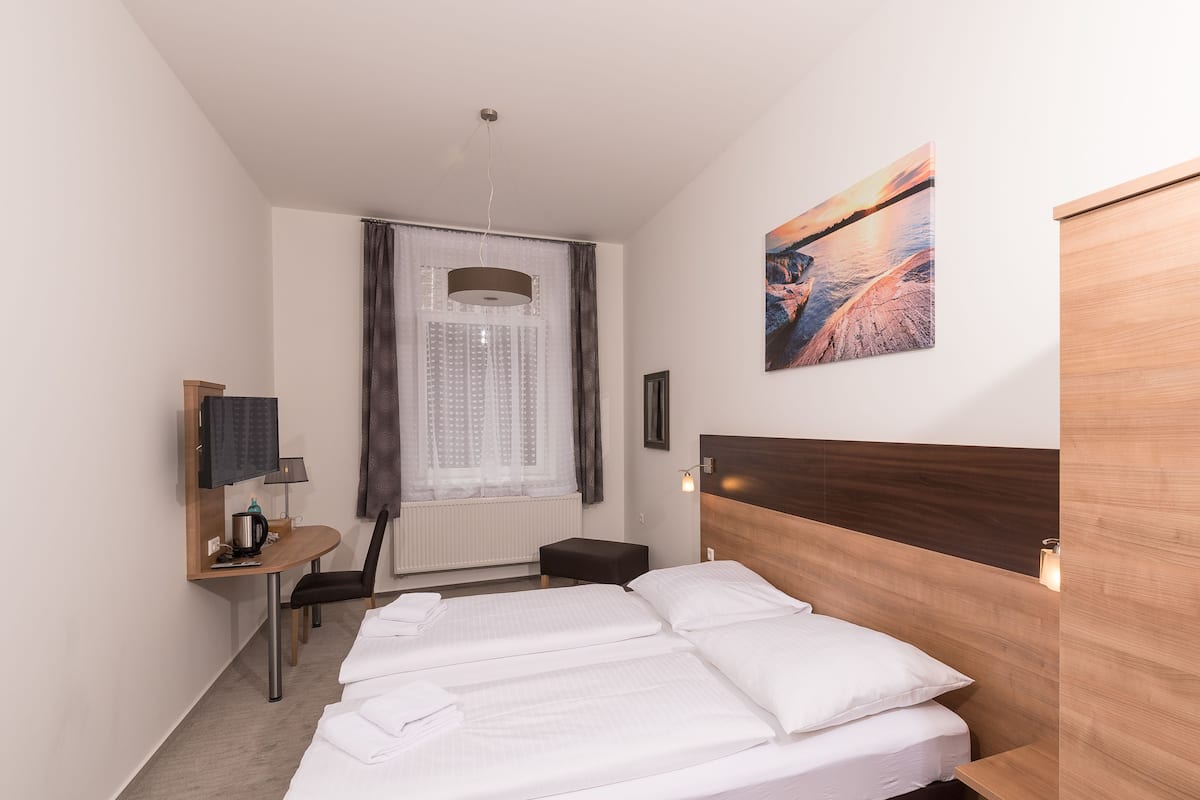 A hotel room with a bed, a desk, a chair, a small table, a radiator, a window with curtains, a painting, and a wall-mounted light fixture.