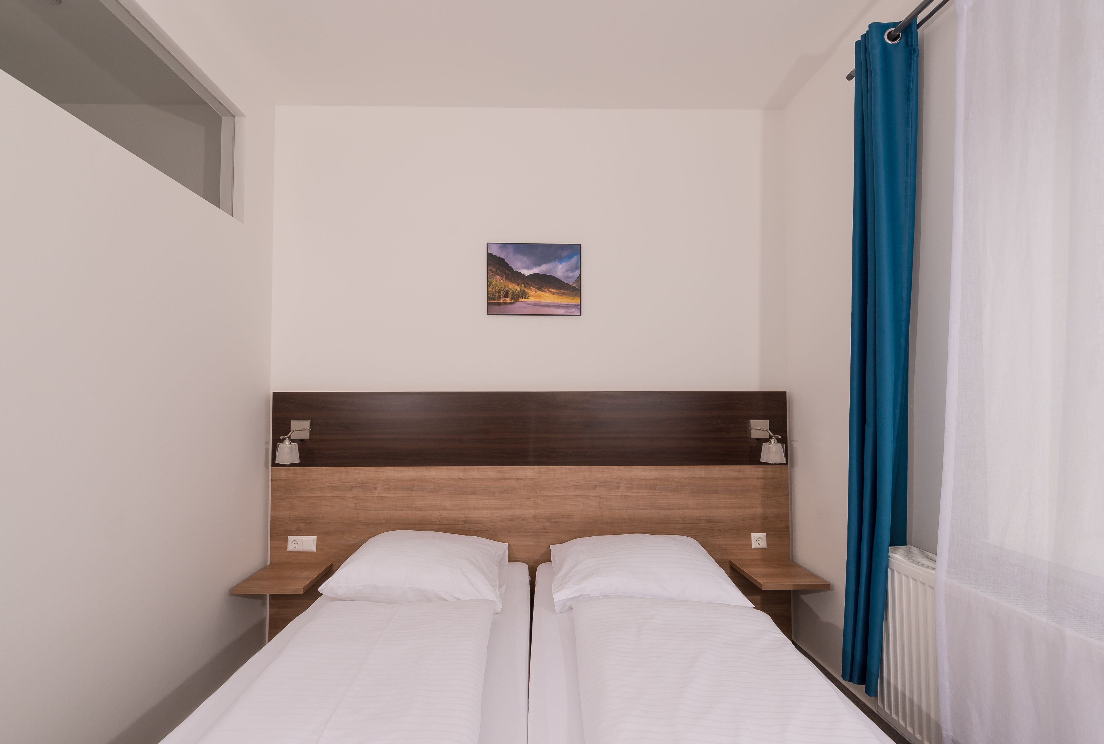 Een tweepersoonsbed met een houten hoofdbord, twee nachtkastjes met lampen, een klein schilderij aan de muur en een raam met blauwe gordijnen.