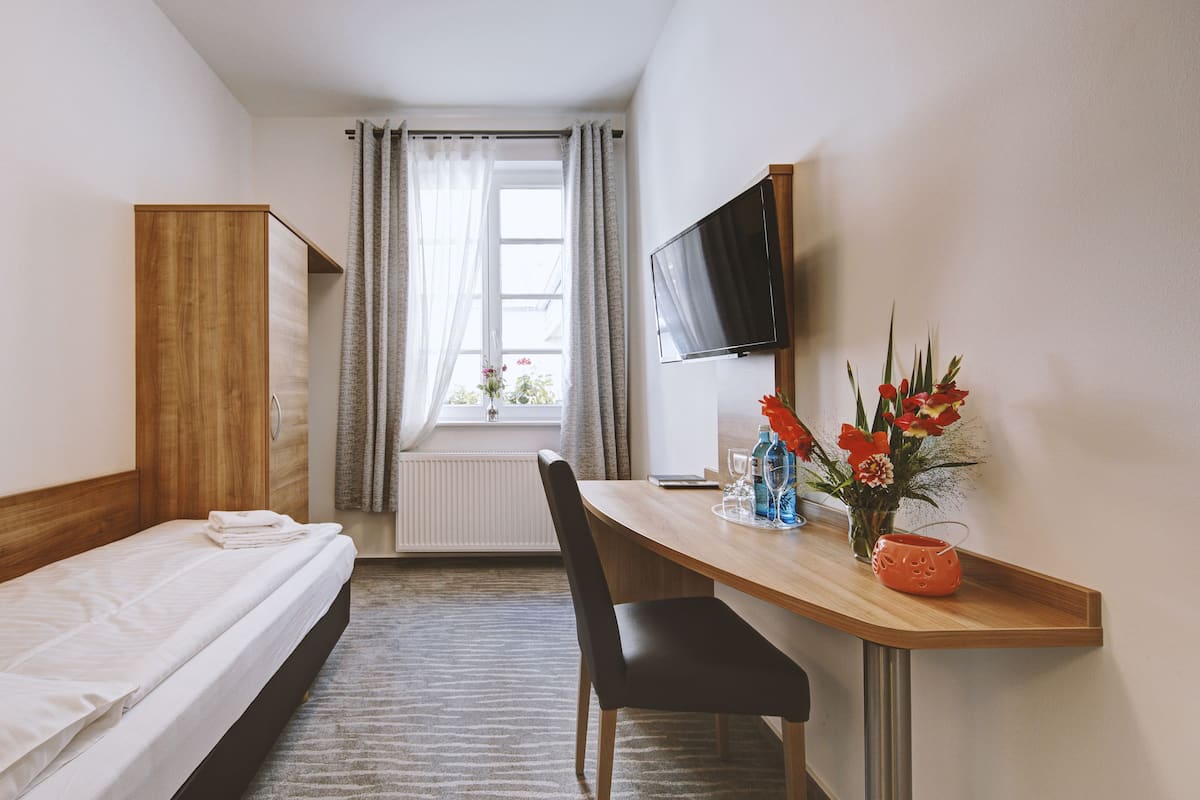 A hotel room with a single bed, a desk with a chair, a television, and a window with curtains.