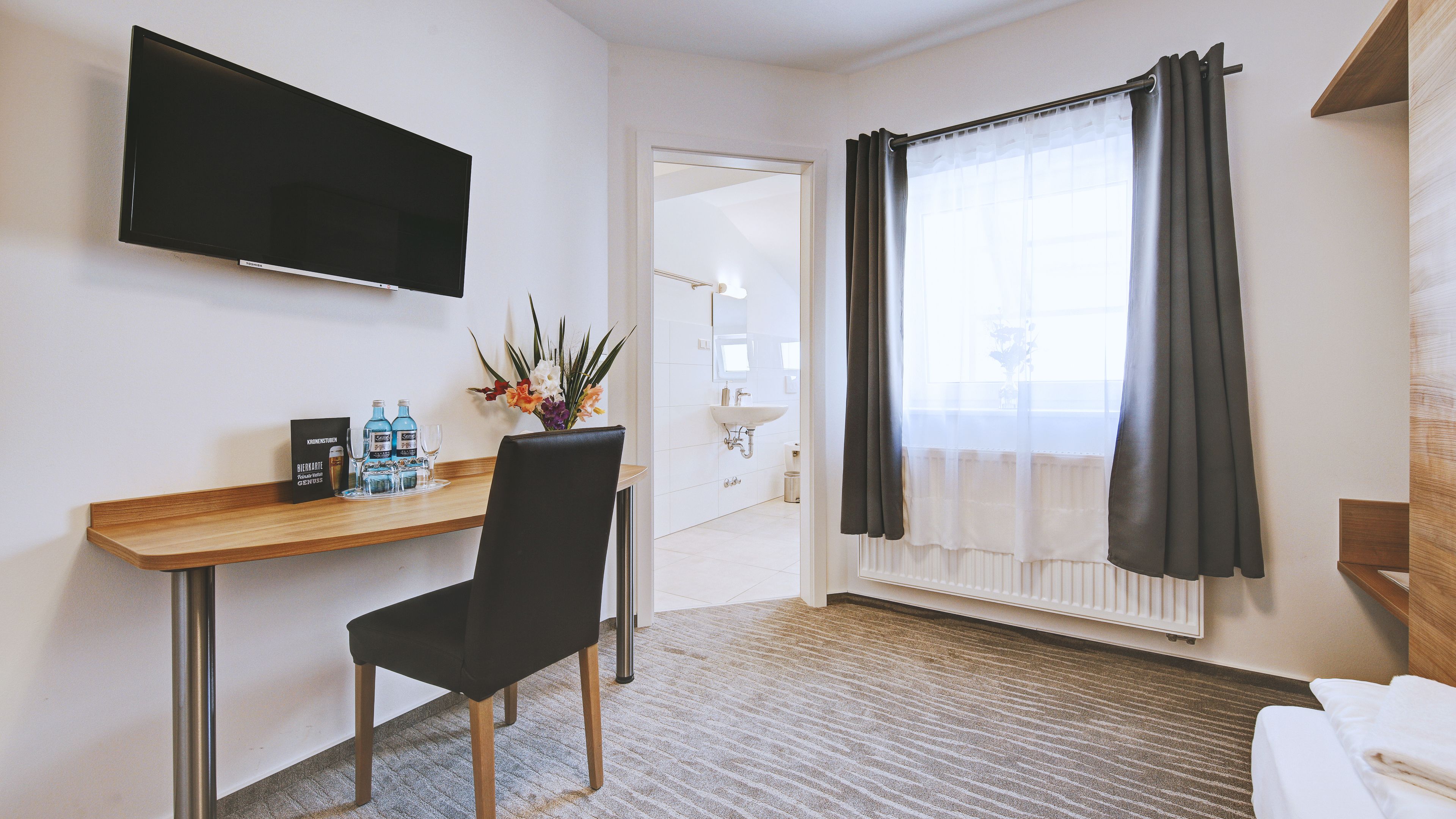 Habitación de hotel con escritorio de madera, una silla negra, televisor de pantalla plana, ventana con cortinas oscuras, lavamanos blanco y una cama.