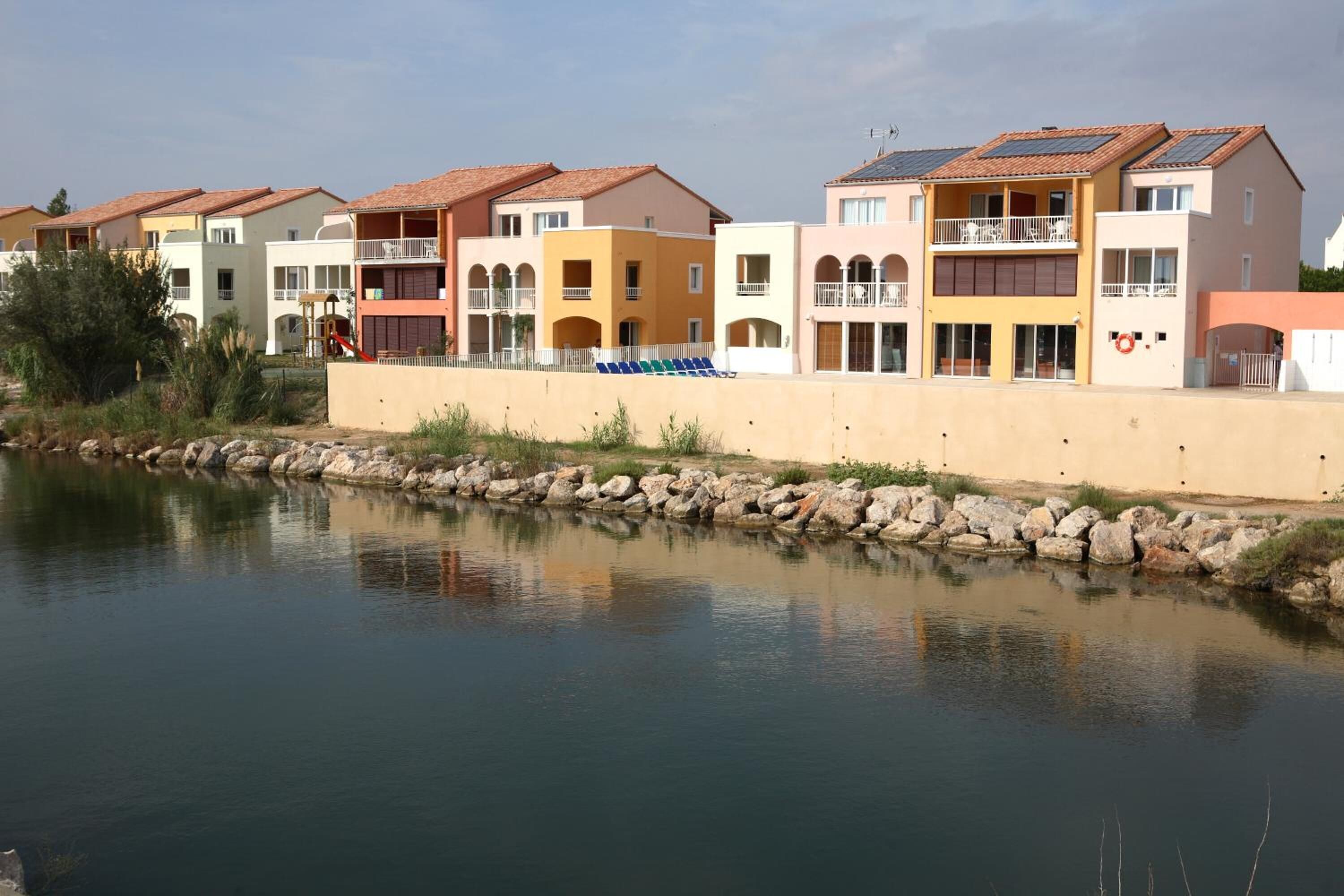 A row of colorful apartment buildings with balconies, a calm water body in the foreground, and a rocky shoreline.