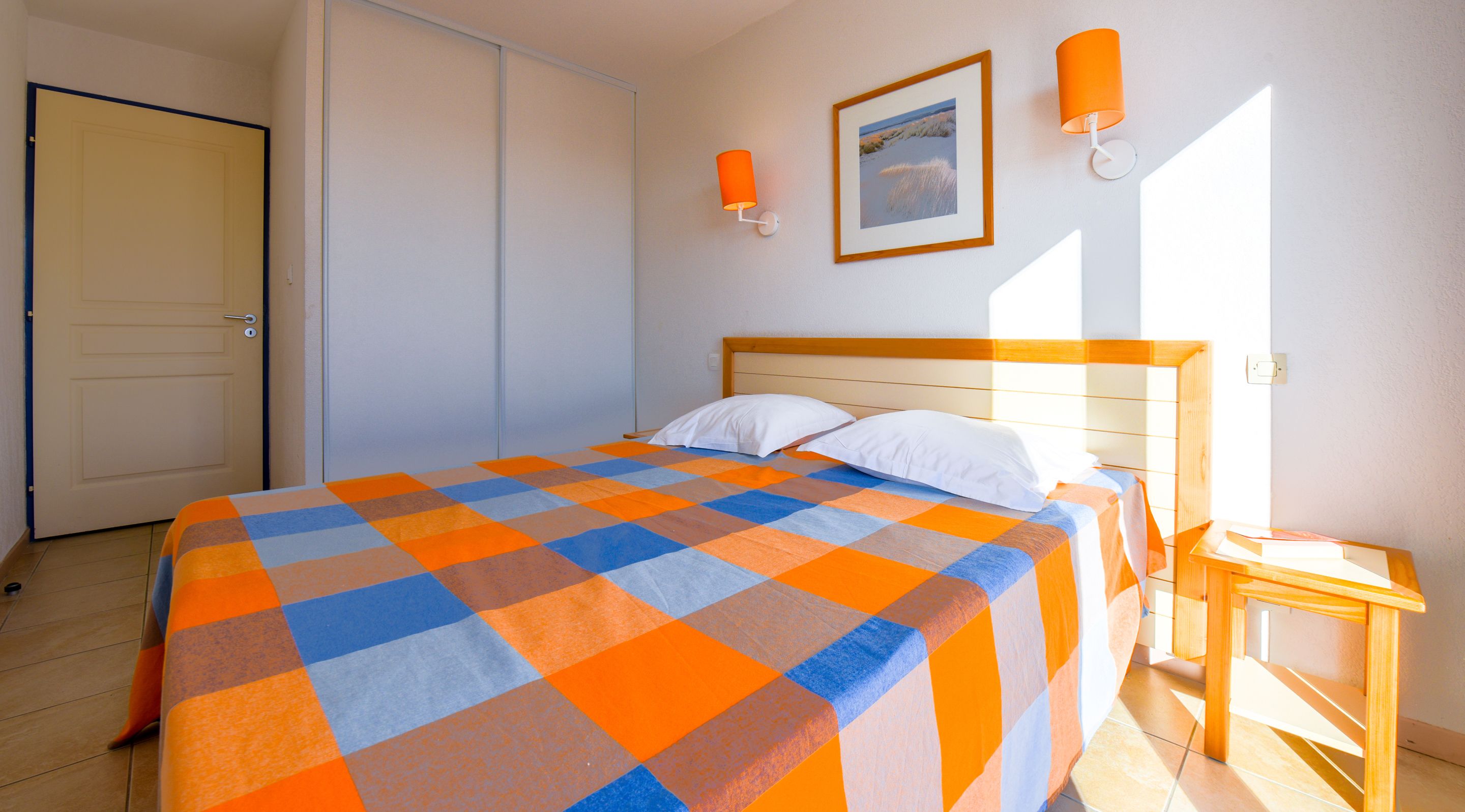 A bedroom with a bed, a nightstand, a framed picture on the wall, and two wall-mounted lamps.
