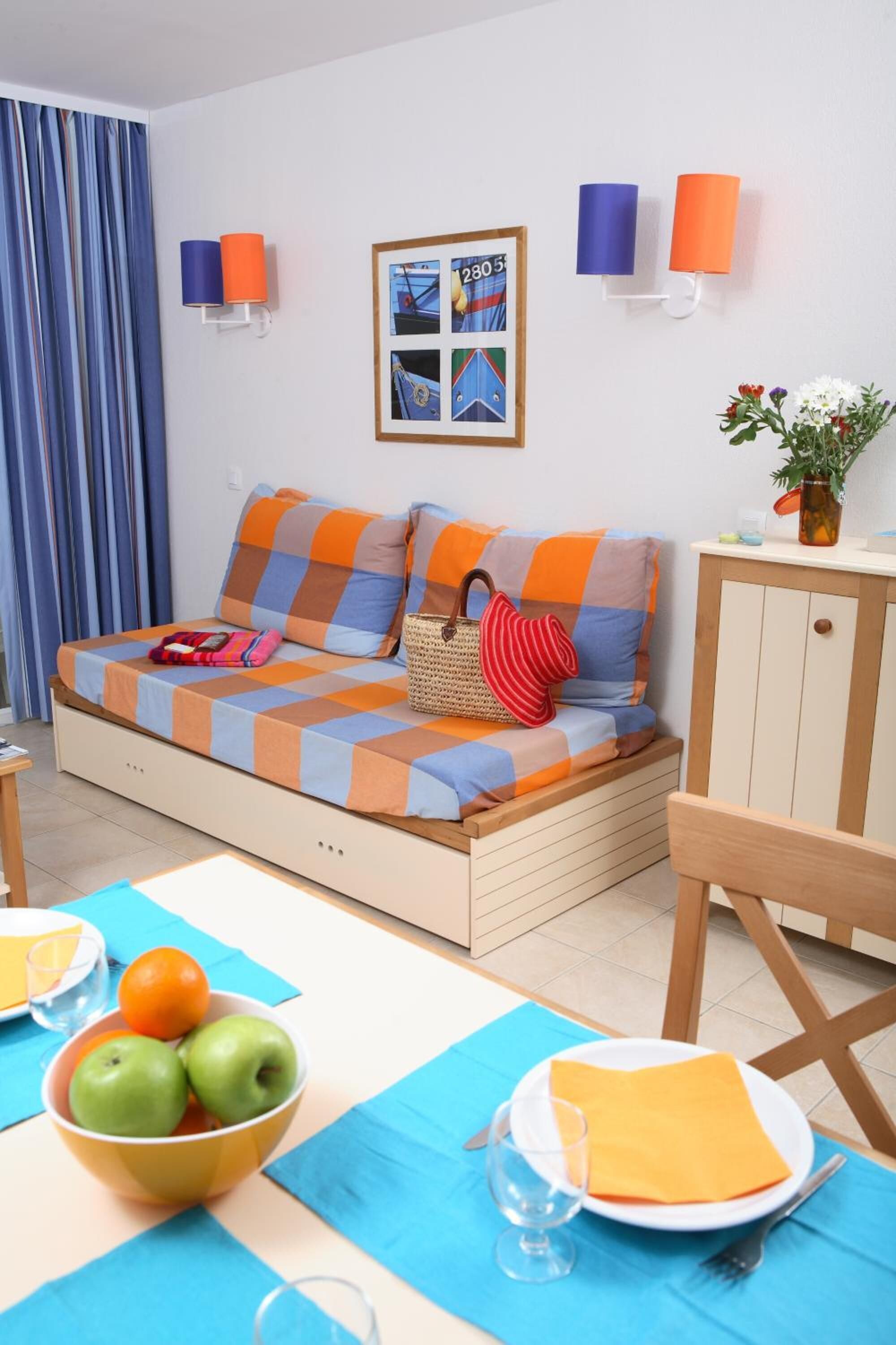 Une pièce confortable avec un lit, une table à manger sur laquelle se trouve un saladier rempli de fruits, et un cadre photo accroché au mur.