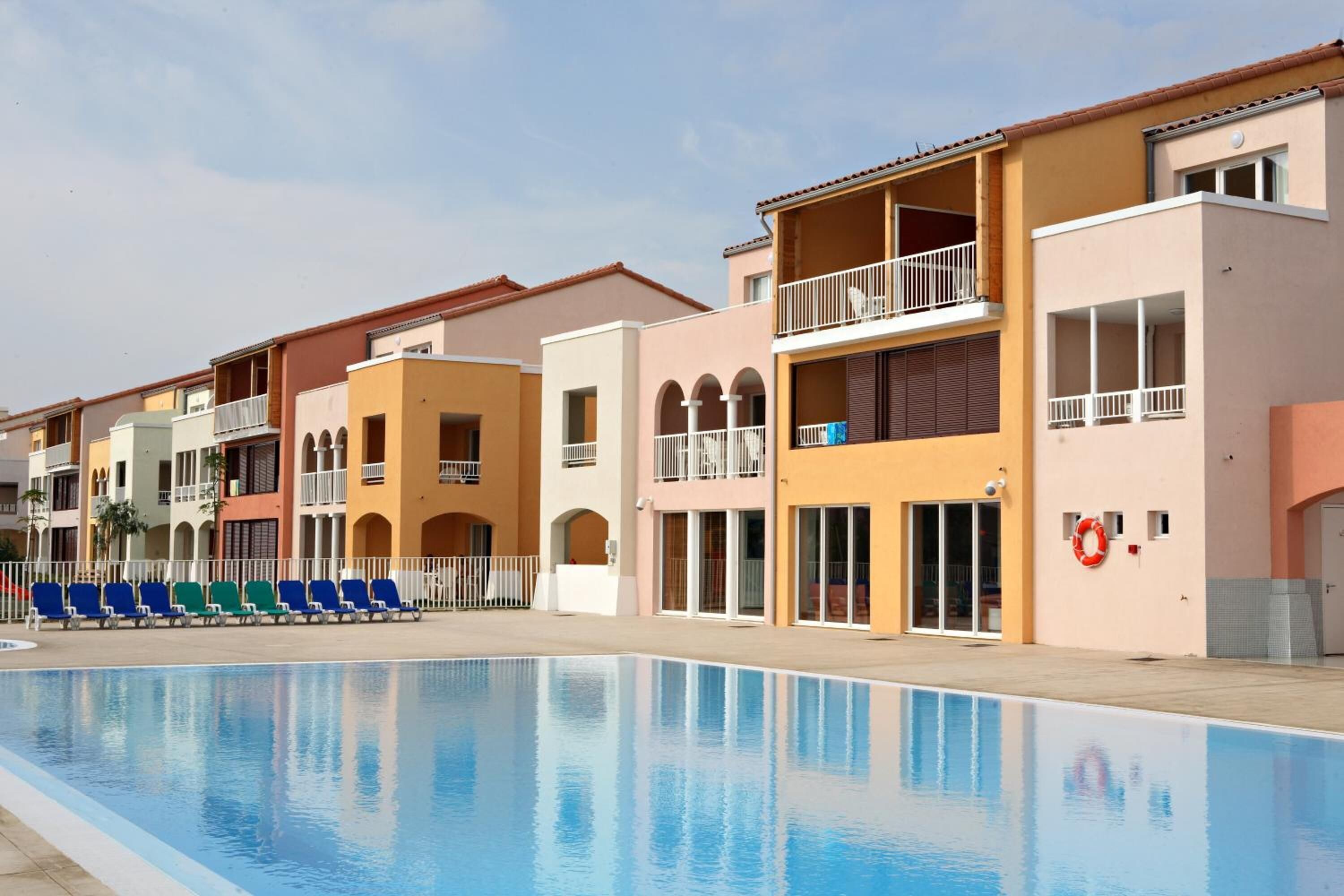 A poolside view of a hotel with multiple balconies, a clear sky, and a row of blue chairs.