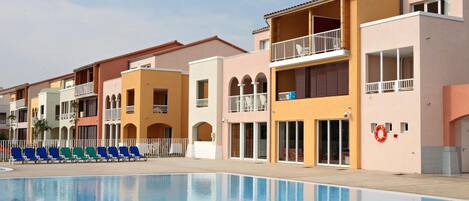 A poolside view of a hotel with multiple balconies, a clear sky, and a row of blue chairs.