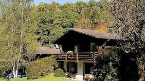 A wooden cabin with a porch, surrounded by lush greenery and trees. - Haus 2 - Typ B (Blockhaus) (Schönecken)