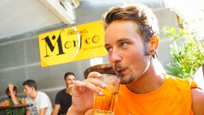 Une personne portant un t-shirt orange avec l'inscription « RIMINI » boit dans un verre. En arrière-plan, on aperçoit des personnes assises à des tables, et une enseigne jaune avec l'inscription « Hôtel Morfeo » est visible.