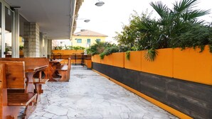 Un espace extérieur aménagé avec des bancs et des tables en bois, un sol carrelé et un mur jaune agrémenté de plantes.