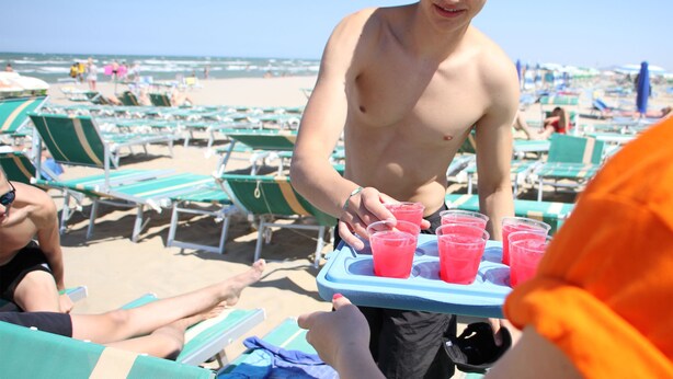 Une scène de plage où l’on voit des gens se détendre sur des transats et une personne qui sert des boissons.