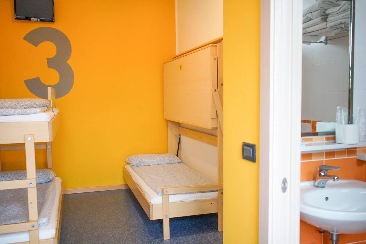 Een kleine, kleurrijke kamer met een stapelbed, een eenpersoonsbed en een badkamer met een wastafel en spiegel.