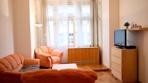 A living room with two orange sofas, a white coffee table, a wooden cabinet with a television, and a window with sheer curtains.