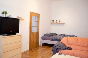 A small, well-lit room with a wooden door, a bed with pillows, a television on a dresser, and a small shelf with plants.