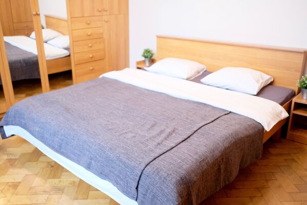 A bed with a grey blanket, wooden headboard, and white pillows. There is a wooden wardrobe with drawers and a small plant on the nightstand.