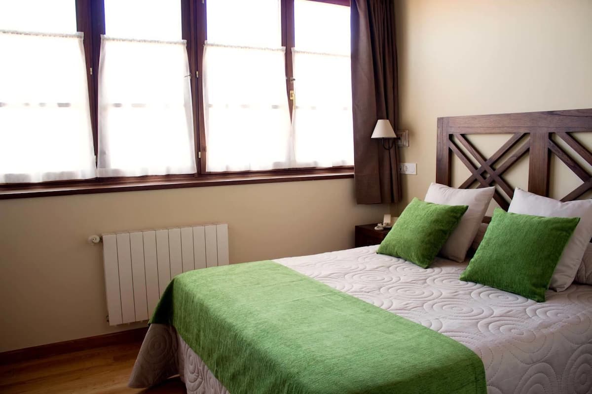 A neatly made bed with green and white bedding, a wooden headboard, a bedside lamp, and a window with curtains.