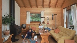 Interior acollidor d'una cabana de fusta amb un sofà, una taula, una llar de foc i una finestra que donen a la vegetació.